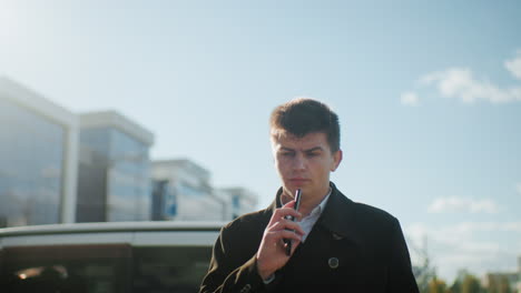 businessman wearing black coat with metallic buttons on phone call outdoors under clear blue sky, looking displeased and contemplative while walking near parked cars in modern urban environment