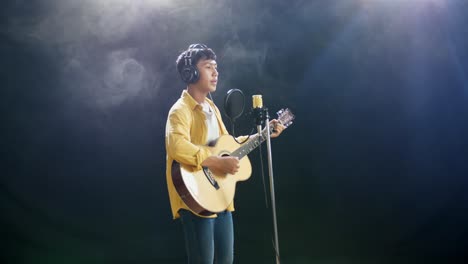 side view of young asian boy with headphone playing a guitar and singing into a condenser microphone on the white smoke black background