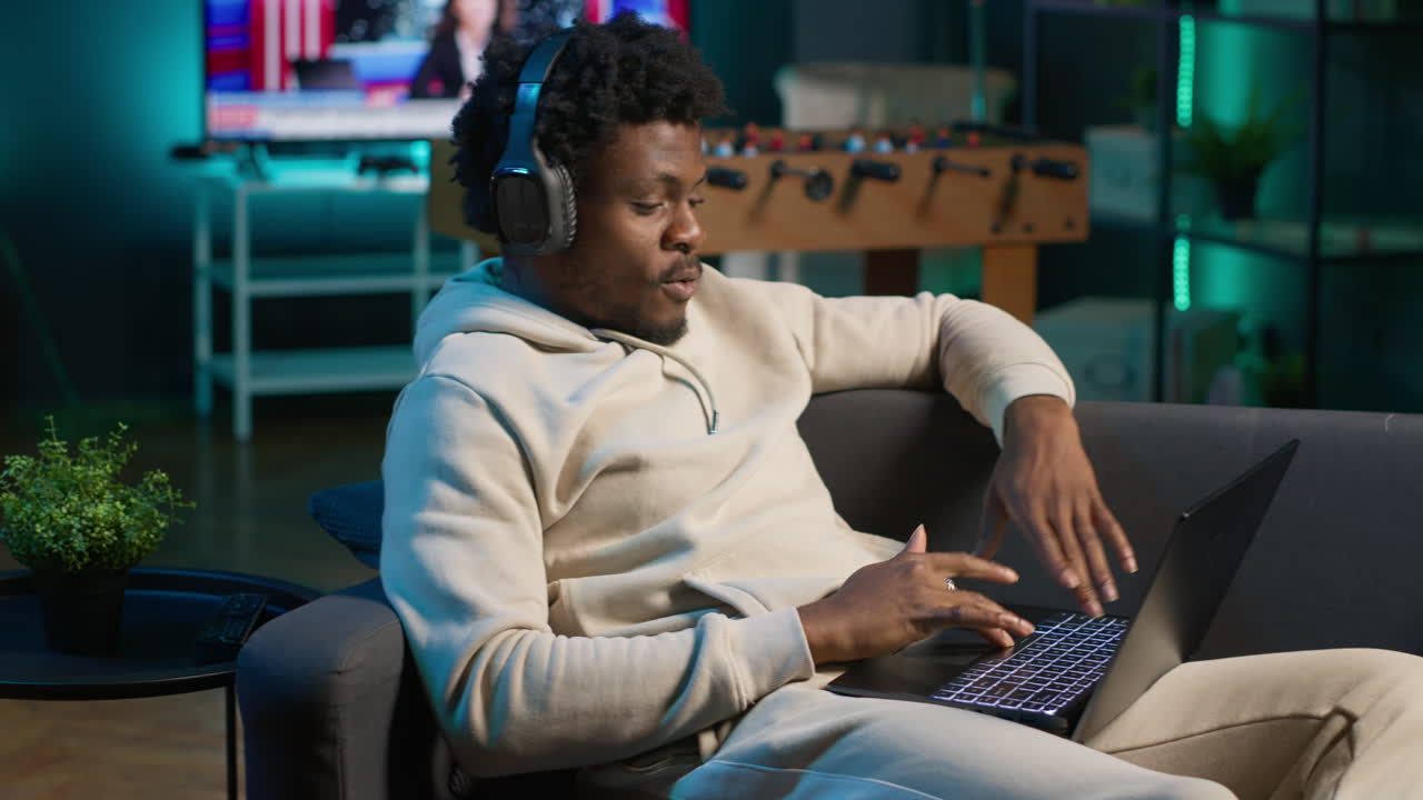 African american self employed man browses the web and listens to music, having fun with songs on headset at his modern home. Young adult laying on couch and relaxing in the living room. Camera A.