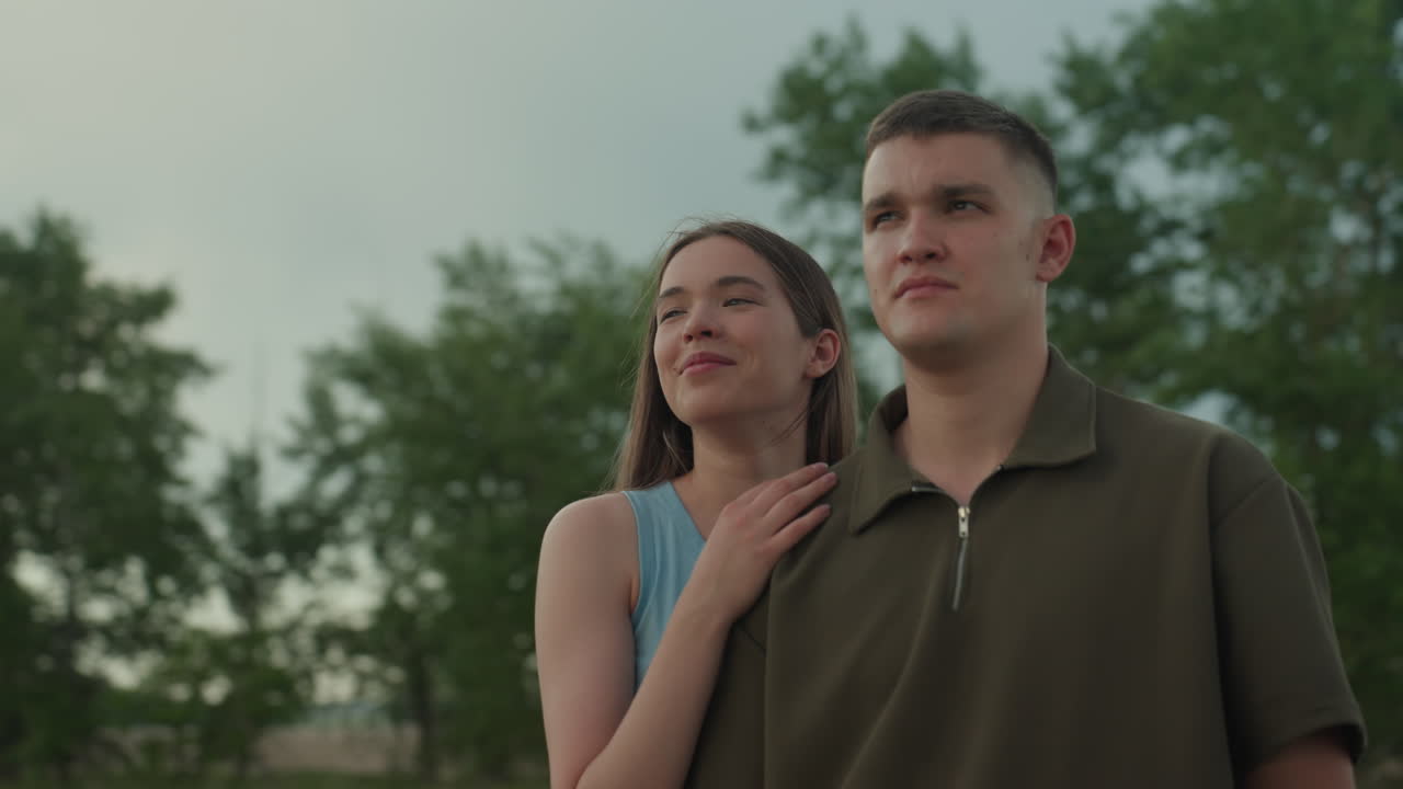 man and woman stand on roadside overlooking expanse of green farm land beneath cloudy sky as woman smiles warmly and places hand on man shoulder