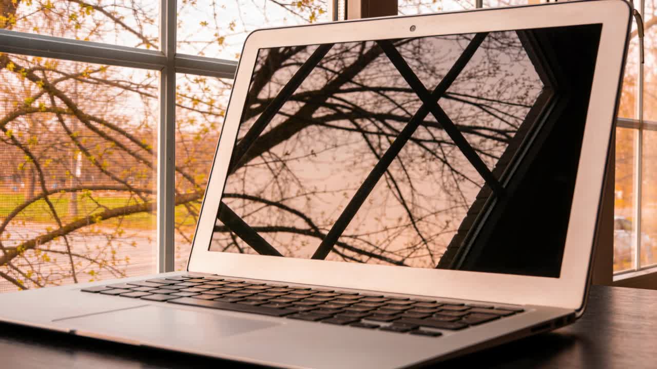 Zoomende kamera indsnævrer billedet på en sølvfarvet bærbar computer ved et vindue, der afslører en detaljeret trærefleksion.