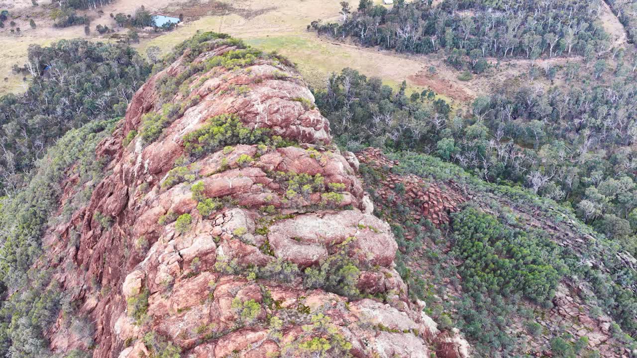 Drone glides above rugged volcanic outcrop, revealing sandstone textures and bushland in soft sunset light