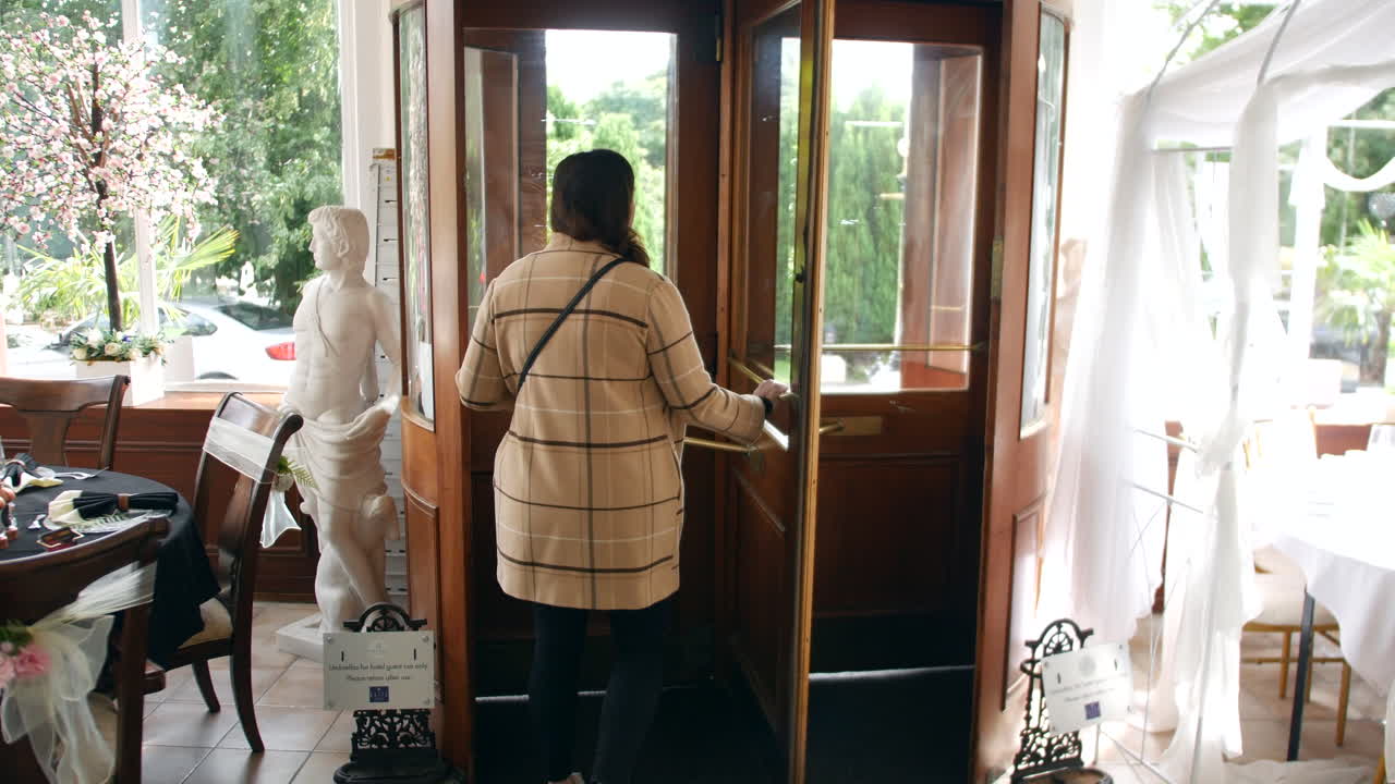 A woman walking through a revolving door, exiting a well-decorated hotel. The room features a statue, floral decorations, and a dining area, with sunlight streaming in through large windows