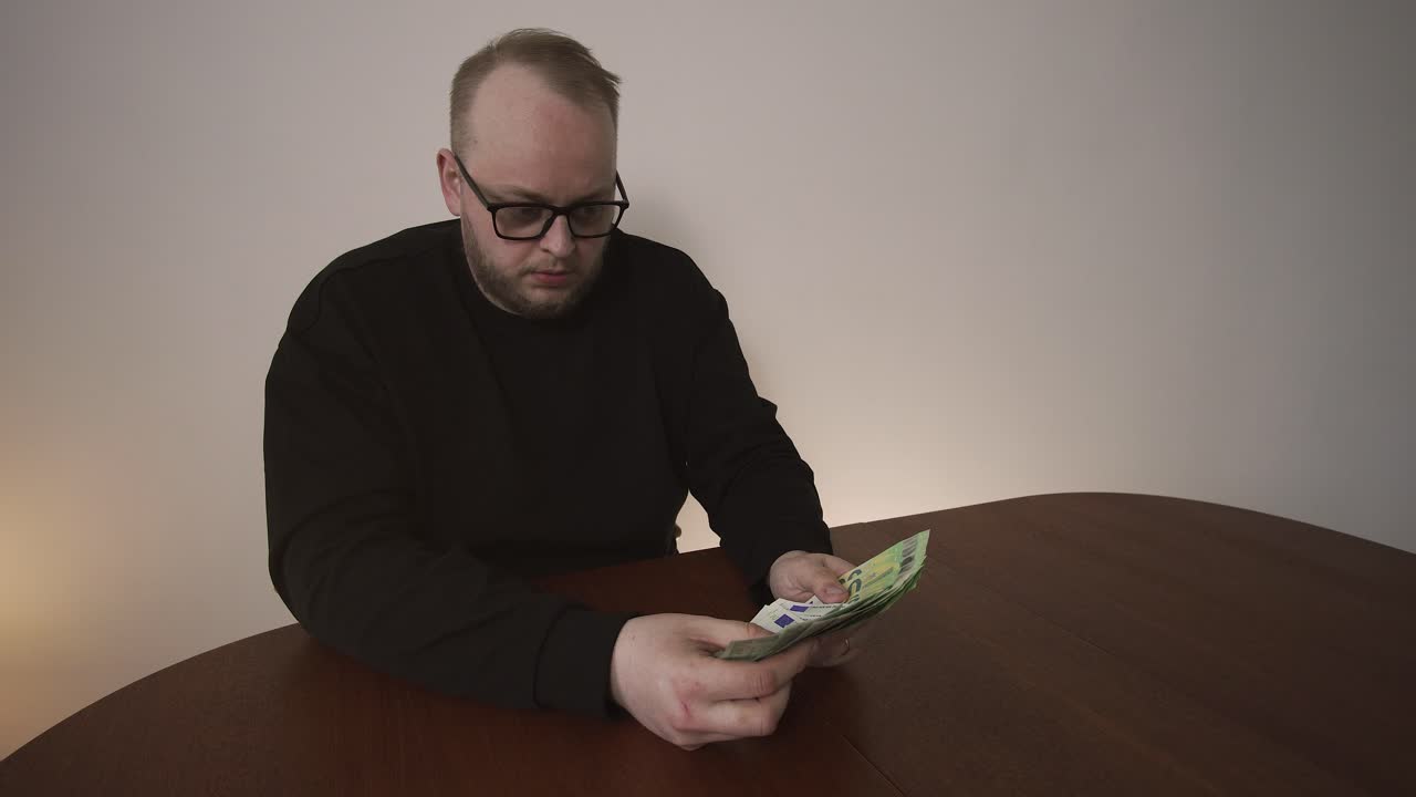 Un uomo sospira deluso mentre conta banconote in euro al tavolo. Concetto di denaro insufficiente, stress finanziario, bisogni insoddisatti e difficoltà di gestione del bilancio.
