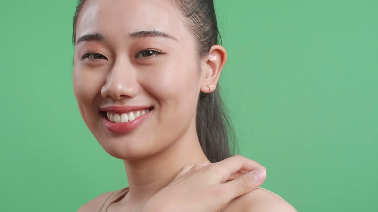 스튜디오에서 녹색 스크린 배경에서 카메라를 바라보면서 피부를 만지고 미소 짓는 아시아 여성의 근접 측면 시각