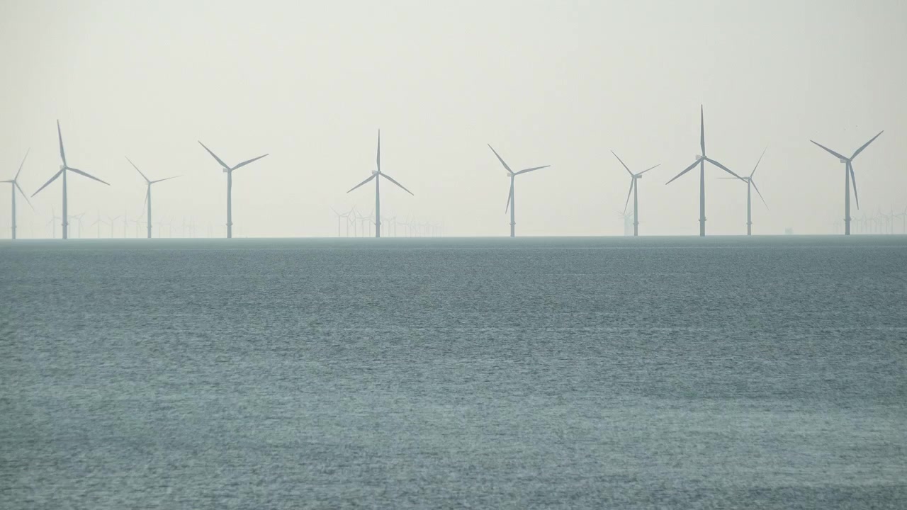 Una fila di turbine eoliche che girano in un grande parco eolico si ergono all'orizzonte molto al largo su un mare calmo.