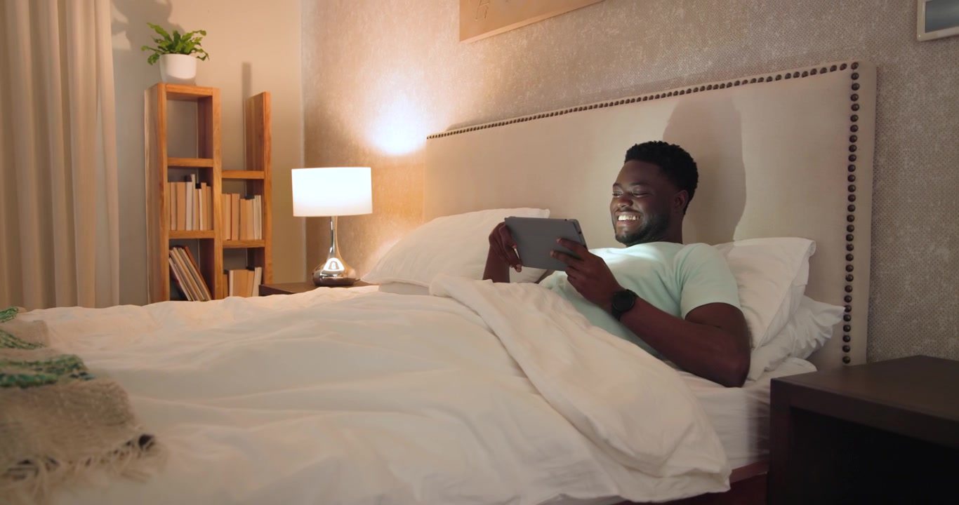 African American man lying in bed laughing while tapping tablet, reacting to funny clip, copy space