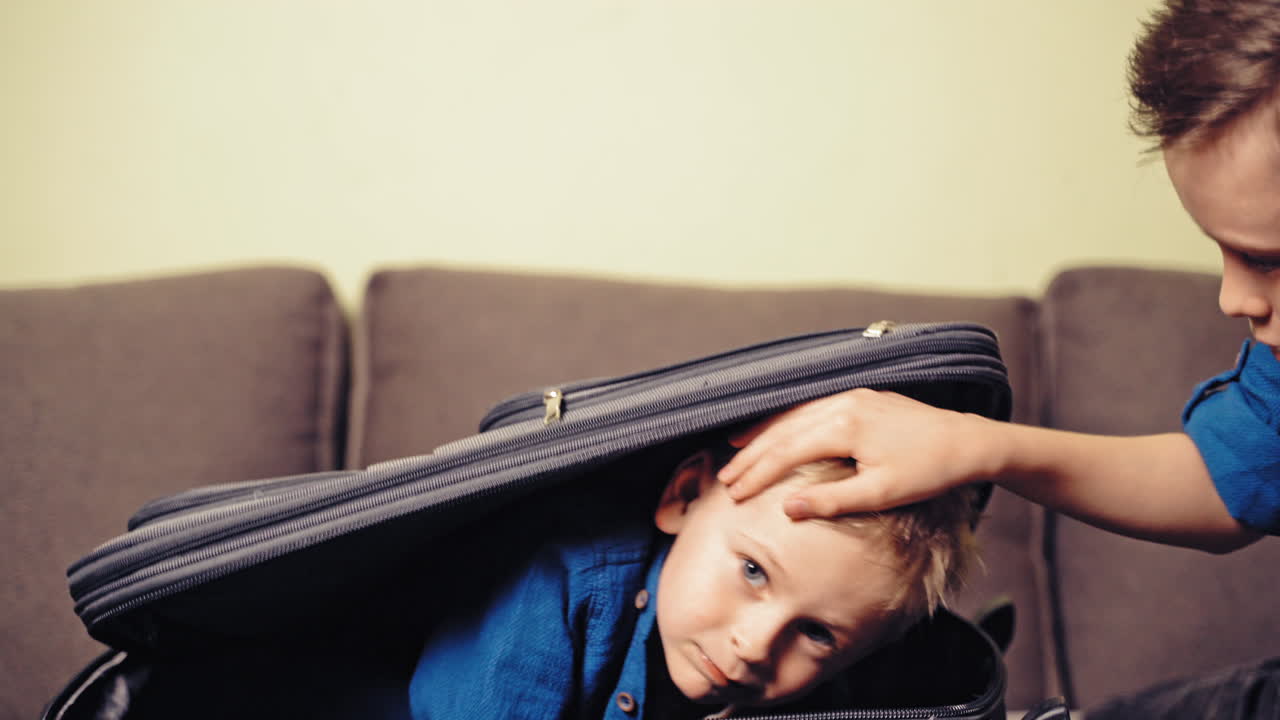 Two cute boys are playing with grey suitcase in the room at home. One little kid is inside the big valise and another boy is closing it.