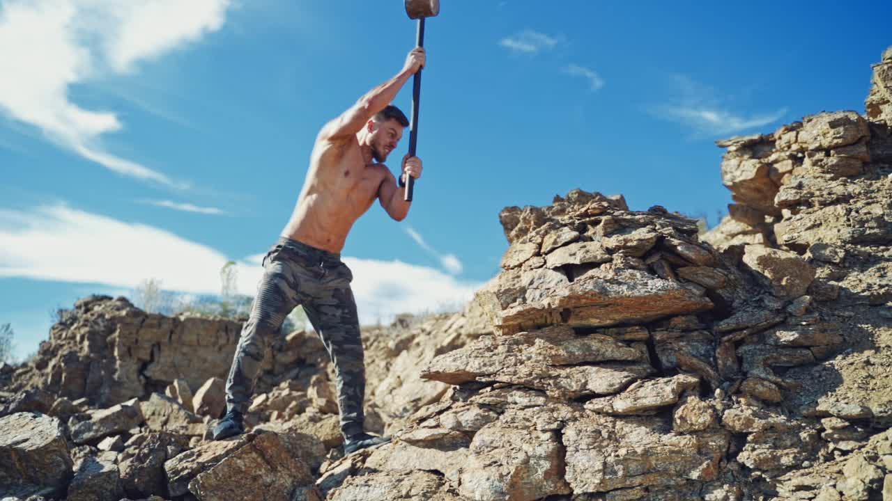 Muscular athlete with hammer on the rocks. Shirtless sportsman breaking stones with hard metal hammer outdoors. Slow motion.