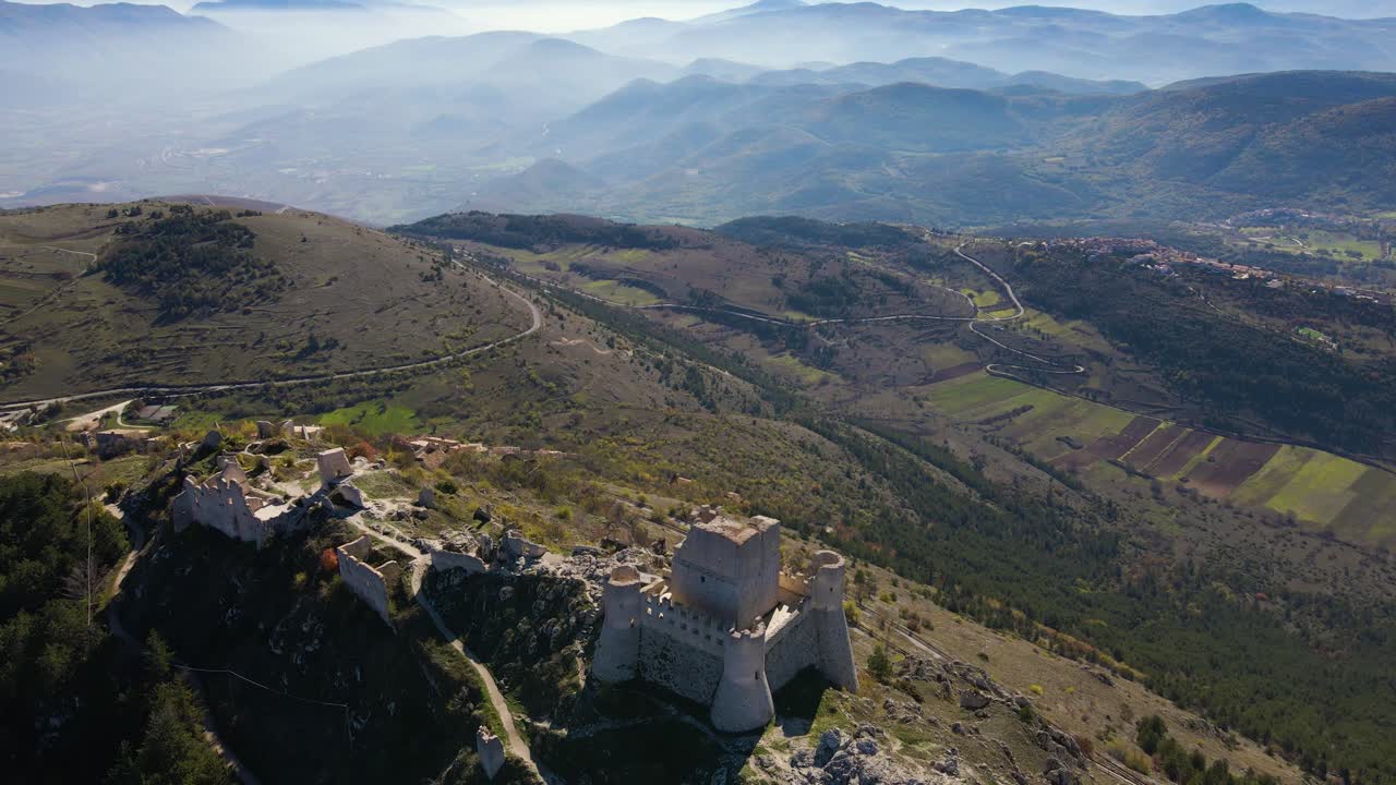 Riprese aeree spettacolari della fortezza di Rocca Calascio su un crinale roccioso, con una nebbia mozzafiato nella valle sottostante, catene montuose stratificate e le suggestive cime del Gran Sasso in lontananza.