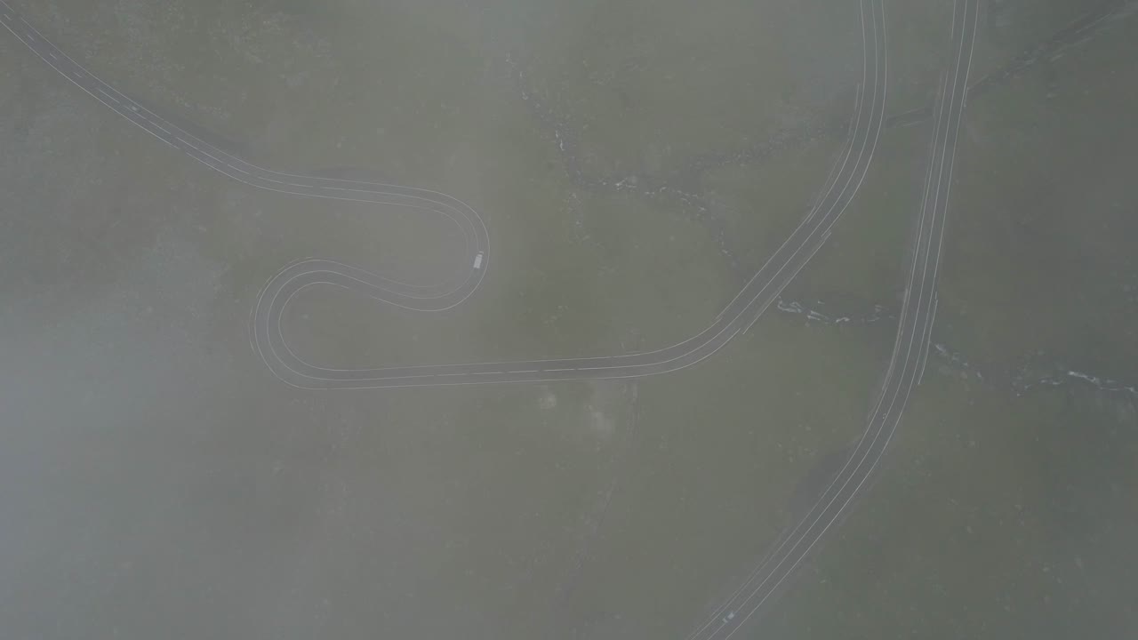 Top-down drone shot descending through white clouds over Transfagarasan road in Romania. A curving mountain route is revealed amid lush green terrain in cinematic 4K motion.
