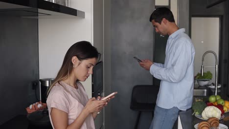 Pareja-Joven-Mirando-Algo-En-La-Pantalla-De-Su-Teléfono-Inteligente-En-La-Cocina-Con-Un-Diseño-Moderno-Tipo-Loft.-Leer-Las-Noticias-De-La-Mañana-O-Consultar-Las-Redes-Sociales