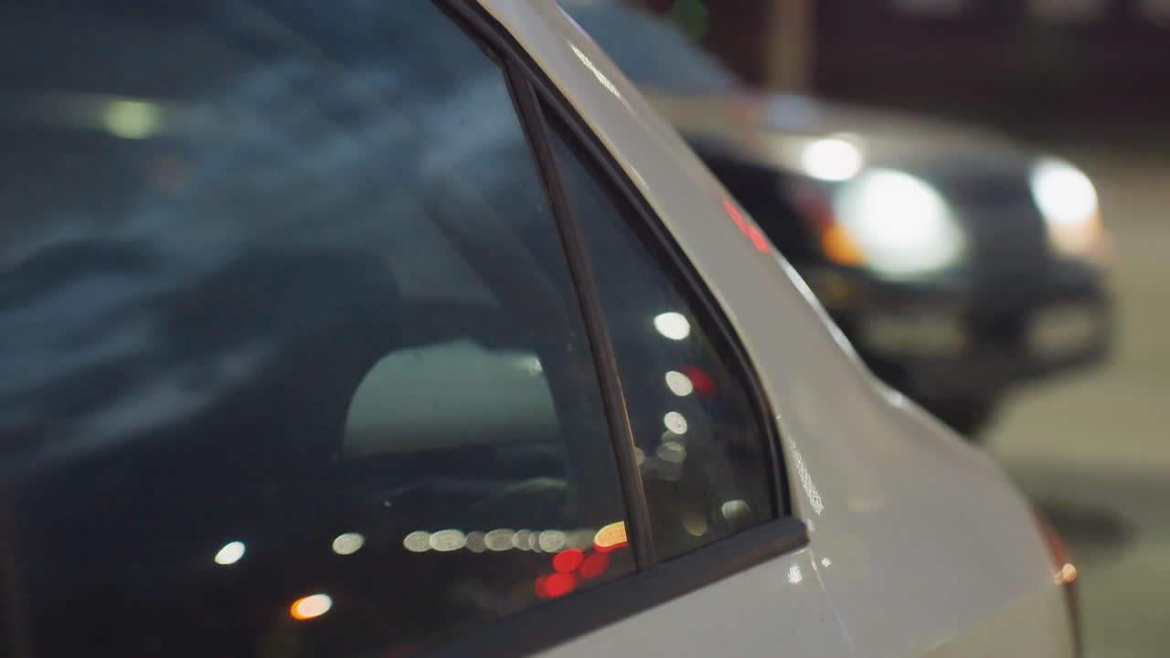 Close up view of parked car side window reflecting blurred city lights and traffic motion, illuminated street scene at night with glowing headlamps, street lights, and people moving in background