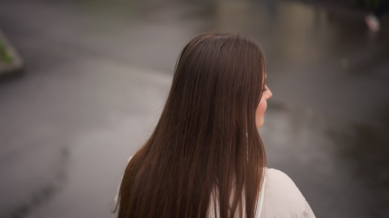 Donna caucasica vista da dietro vicino a una pozzanghera, lunghi capelli lisci che ricadono su un cappotto chiaro, riflesso della strada bagnata e toni spenti, cielo nuvoloso che crea una luce soffusa, solitudine contemplativa e osservazione silenziosa