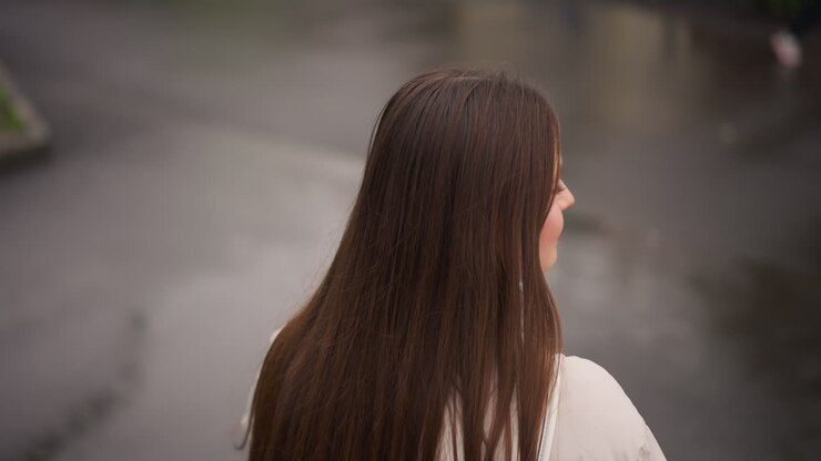 Mujer caucásica de espaldas junto a un charco, pelo largo y liso cayendo sobre un abrigo claro, reflejo de la calle mojada y tonos apagados, cielo cubierto creando una luz suave, contemplativa soledad y silenciosa observación.