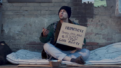 homeless man sitting on street, holding sign and pleading for money