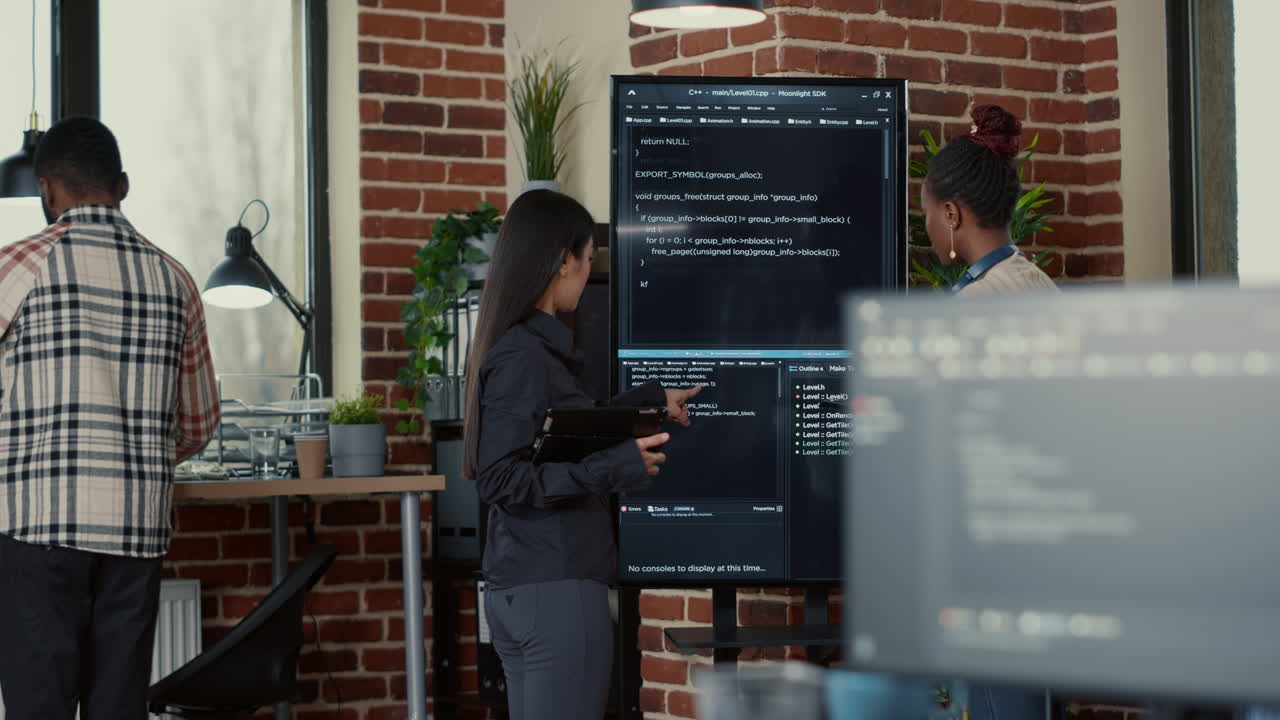 Software engineer holding digital tablet analyzing code on wall screen tv explaining errors to colleague programer