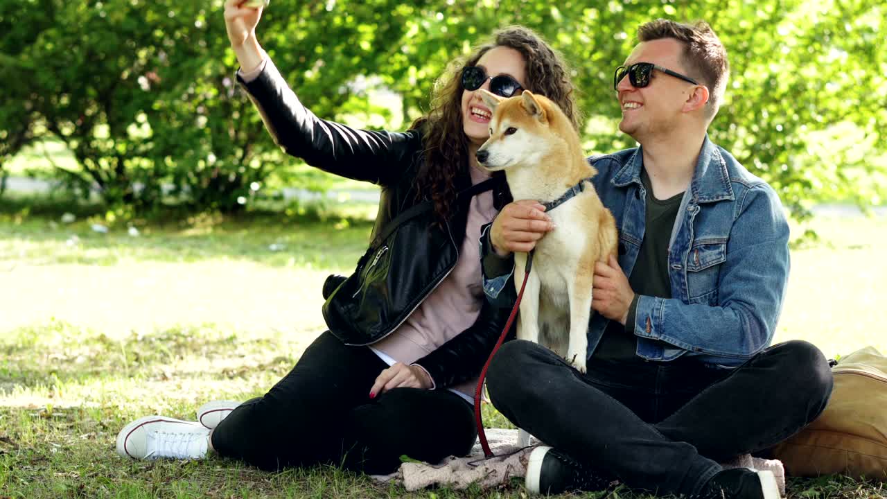 행복한 부부는 스마트폰을 사용하여 개와 셀피를 찍고 공원의 잔디에 앉아 있습니다. 사람들이 포즈를 취하고, 이야기하고, 시바 이누 강아지를 만지고 있습니다.