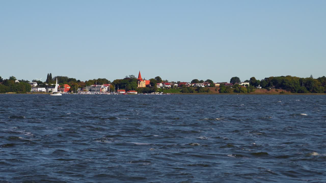 small village across coast sea with sailboat crossing left to right