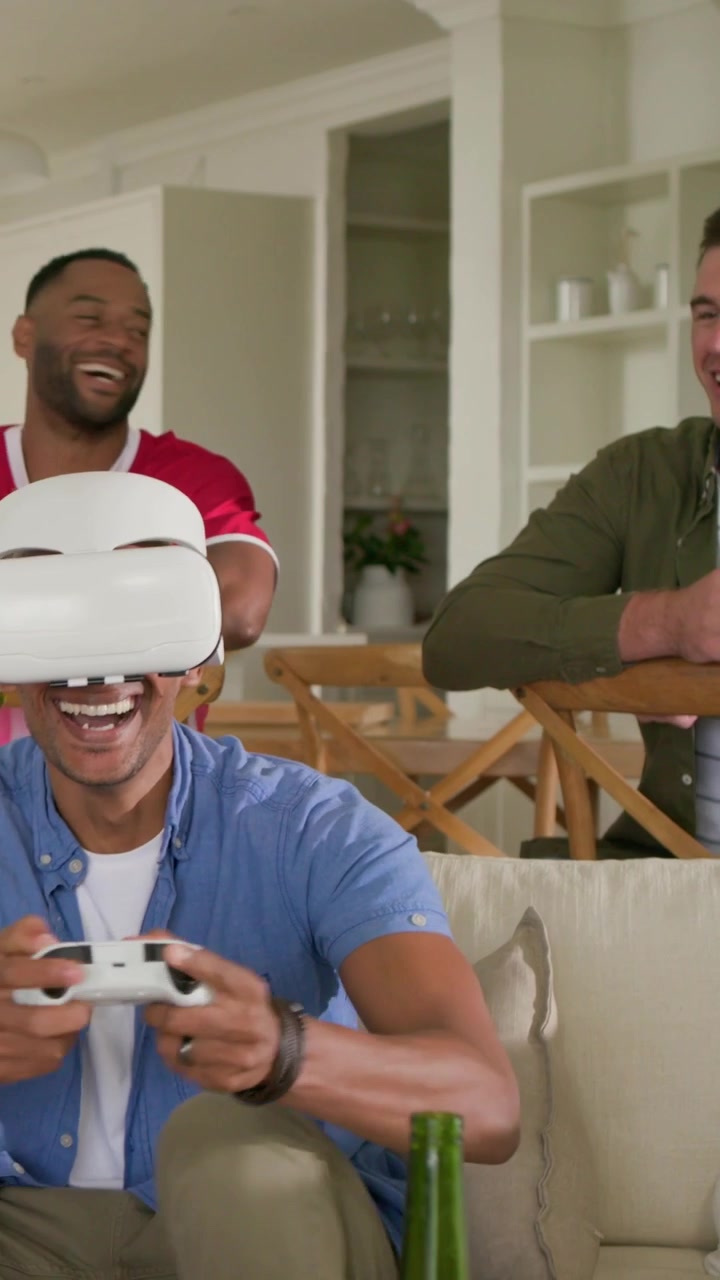 Video verticale: Uomo che indossa un visore VR, in camicia blu, usa un controller a casa, amici che fanno il tifo