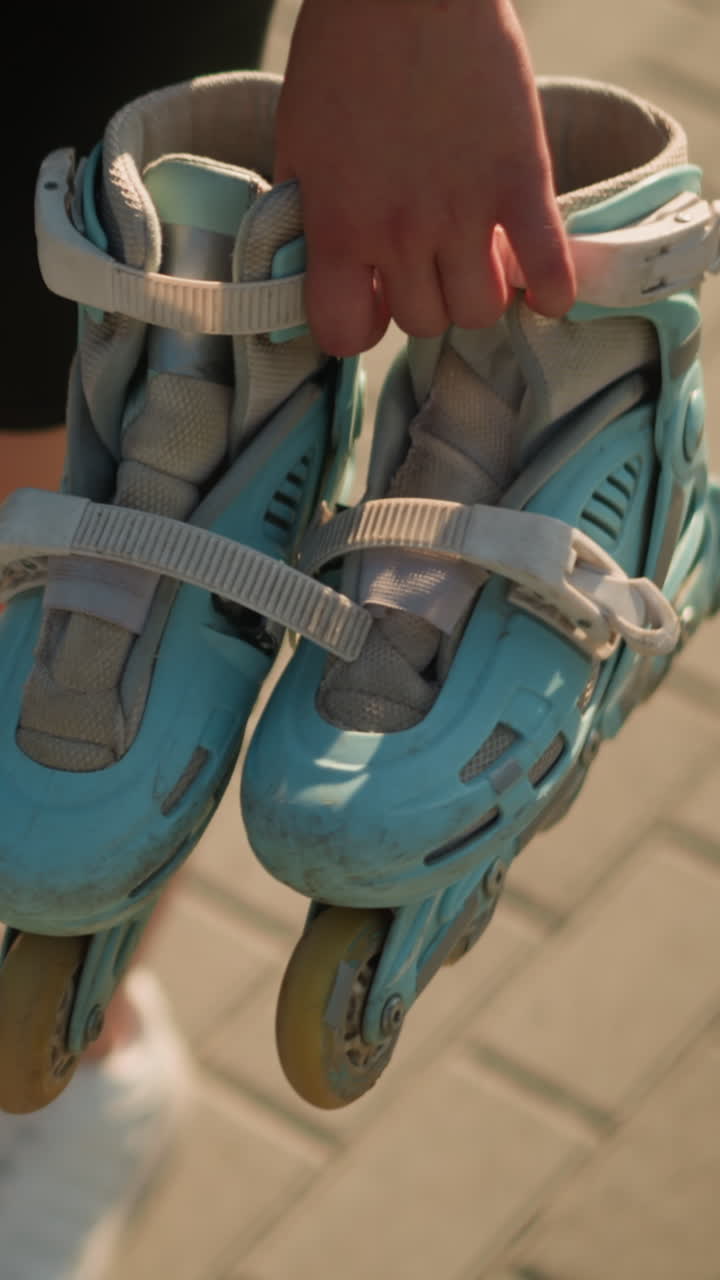 Young lady wearing black tights walking outdoors holding cyan roller skates in her right hand, visible details include a black wristband and an interlocked path background