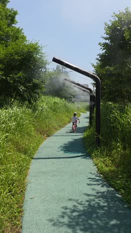a little girl, seen from behind, rides her bicycle with training wheels through a cooling water mist system designed to combat the heat on a public path in yangjaecheon park, seoul on hot summer day