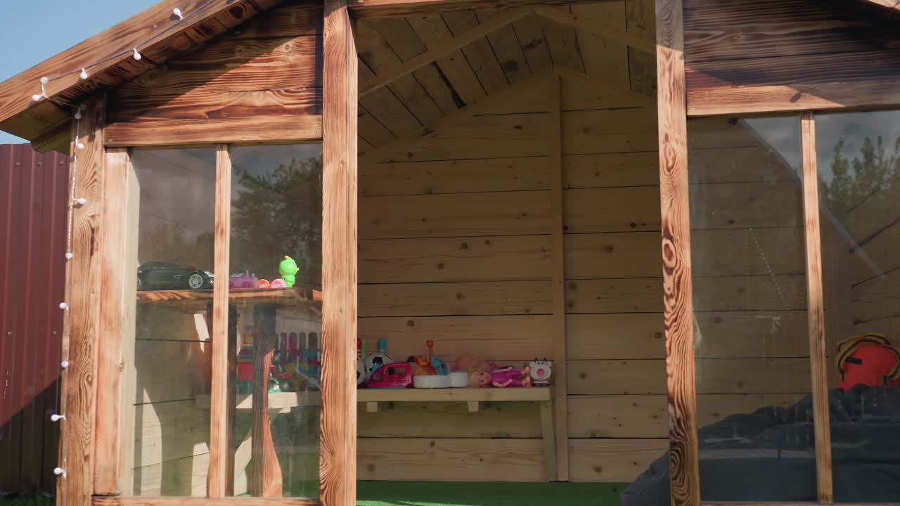 Wooden kid playhouse with glass panels standing on green grass in sunny backyard, surrounded by red metal fence, tall pine tree, and adjacent sandbox filled with outdoor toys and bright lighting