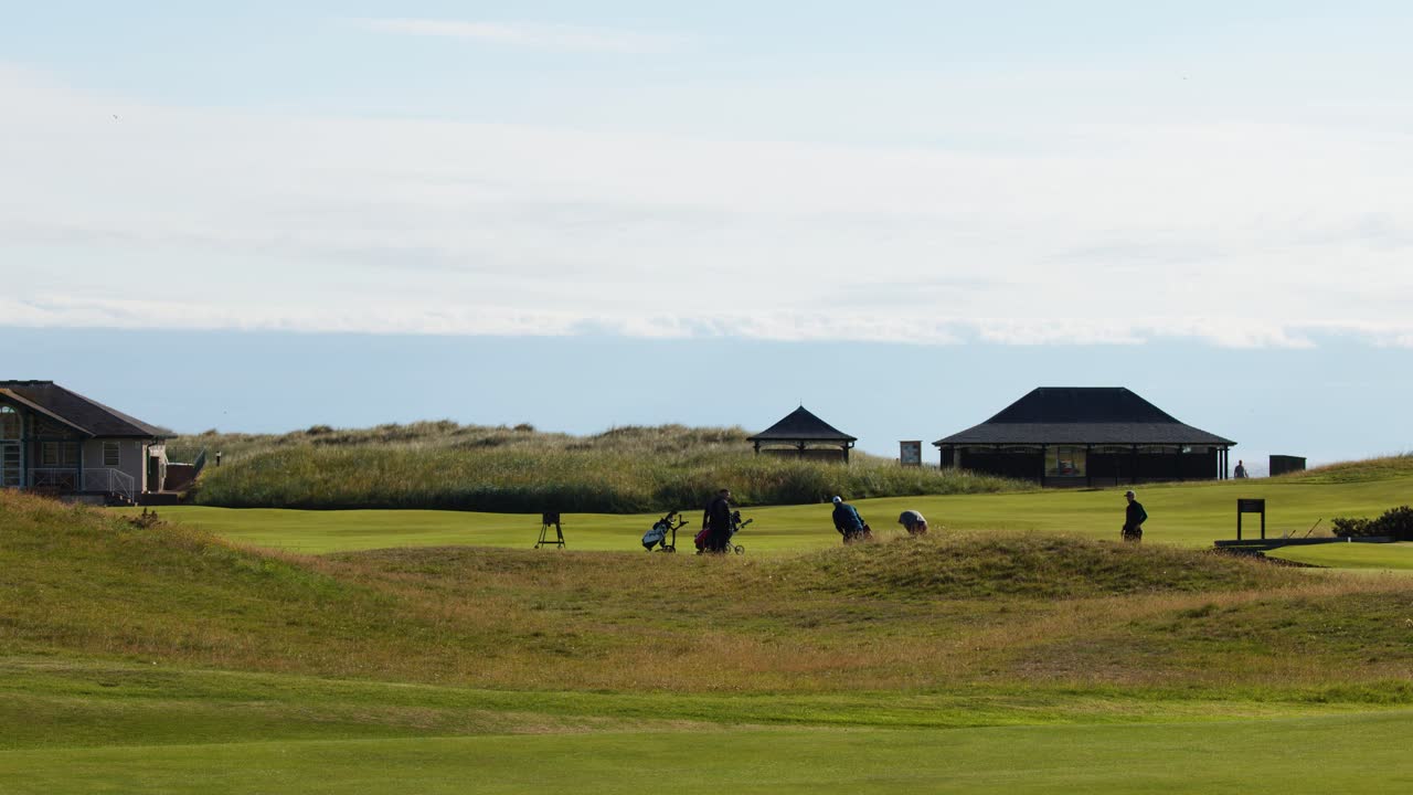 Golfers walk and play on historic St Andrews course, bright daylight, wide static camera shot