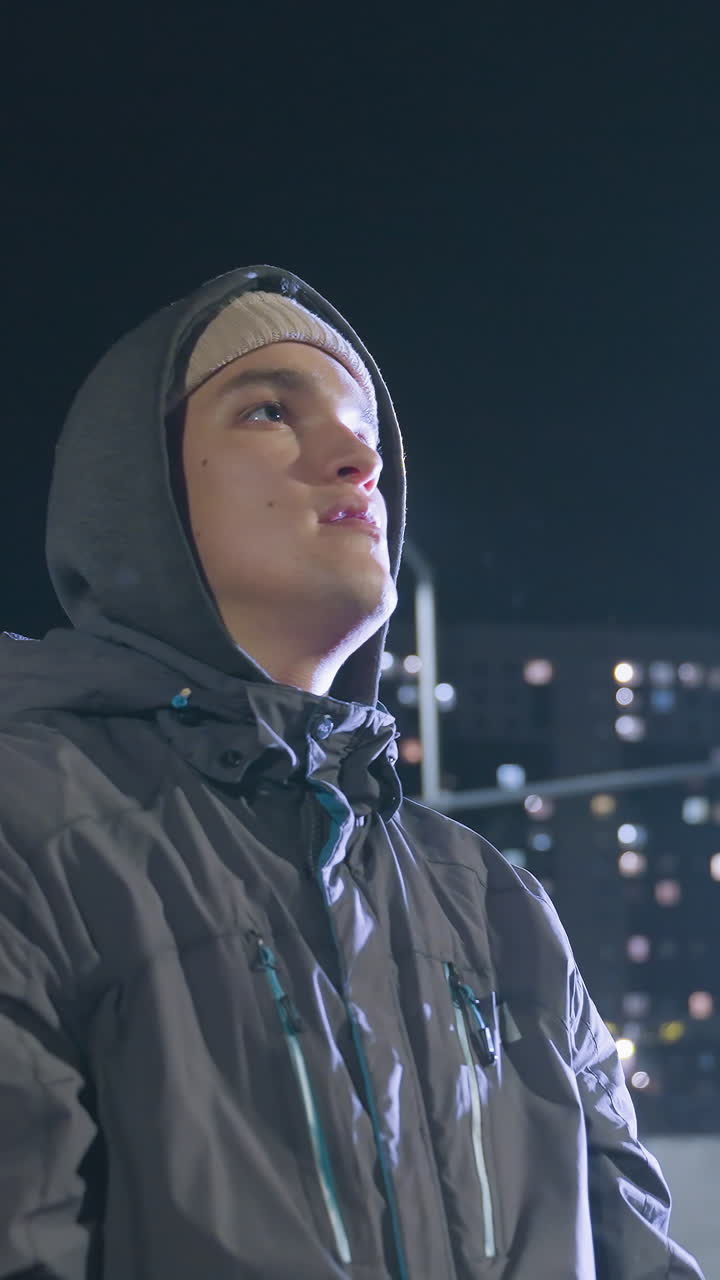 vista lateral de un futbolista con guantes lanzando una pelota de fútbol al aire y atrapándola durante una sesión de entrenamiento nocturna en un campo urbano, luces brillantes de la ciudad y un fondo borroso