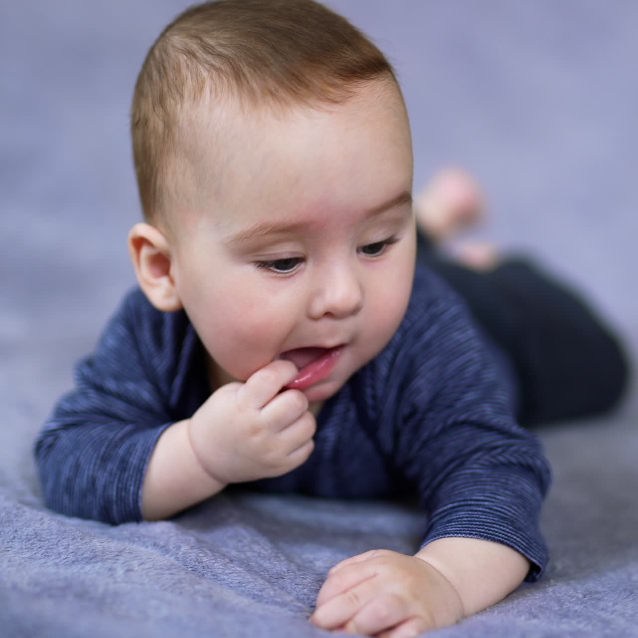 Adorable baby boy pulls his lower lip while lies on bed. Sweet little toddler chews his finger and looks around. Grey blurred backdrop