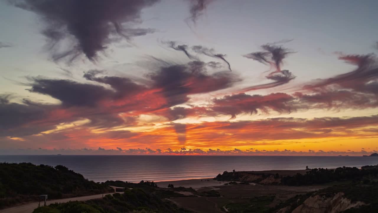 Die vom Sonnenuntergang beleuchteten Wolken verdunkeln sich, während die Sonne an einer Klippe über dem Meer versinkt und Dünen sowie Dämmerungsstrahlen färbt.