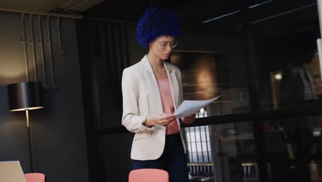 mujer de negocios casual biracial con afro azul sosteniendo documentos y usando computadora portátil, cámara lenta