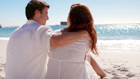 Happy-couple-sitting-together-on-the-sand-looking-at-each-other