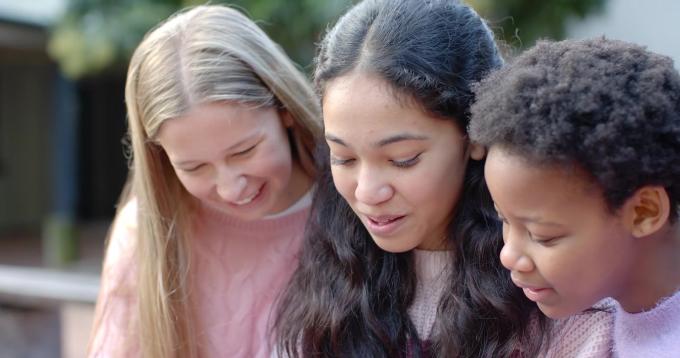 Diverse tenåringsjenter ser på noe utenfor bildet, lener seg, smiler og fokuserer på en utebenk i gensere.