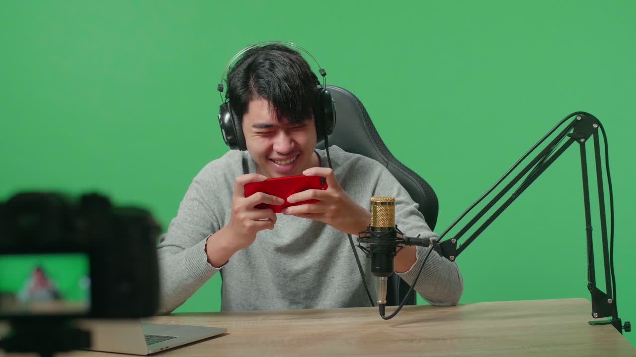 un hombre asiático sonriente con auriculares y computadora jugando a un juego de teléfono móvil mientras graba video con la cámara en la pantalla verde
