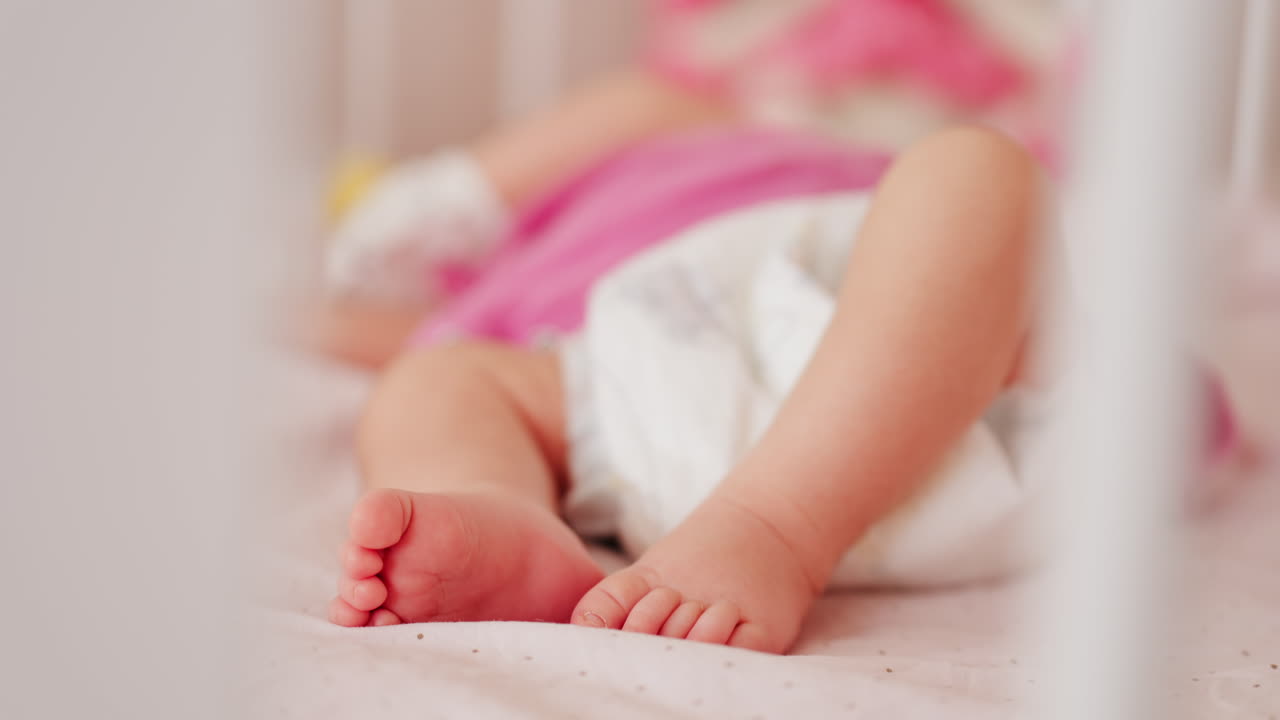 Close up of a baby's legs and diaper as the infant rests in a crib, seen through the crib bars