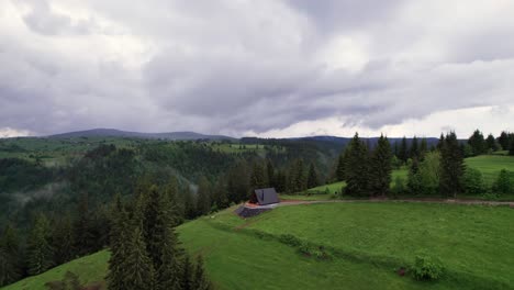 Aerial-view-of-a-remote-A-frame-cabin-in-a-evergreen-forest,-alpine-background