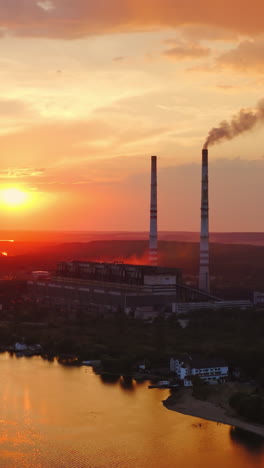 industry near the river at setting time. massive smoke from industrial chimney on pink sky background in the evening. harmful factory in the countryside. aerial view. vertical video