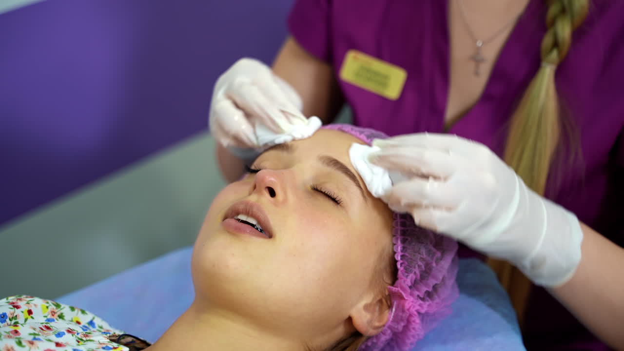 Cosmetologist cleans the skin of a woman's face before a cosmetic procedure by using napkins. Video from the top.