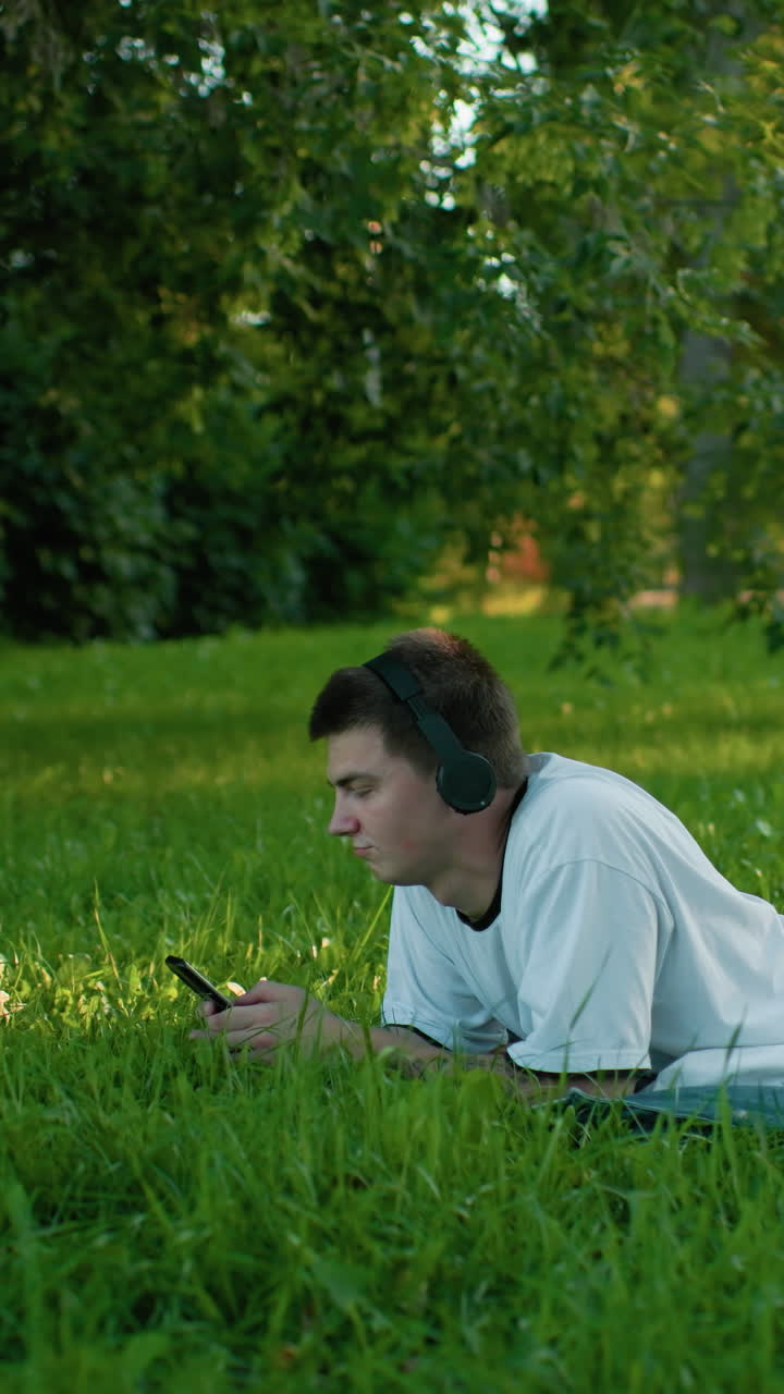 잔디 에 매트에 누워있는 젊은 남자의 측면 뷰 헤드폰을 착용, 그의 전화에 초점을 맞추고, 따뜻한 빛 아래 나무로 둘러싸인 평화로운 녹색 공원에서 음악에 머리를 흔들고