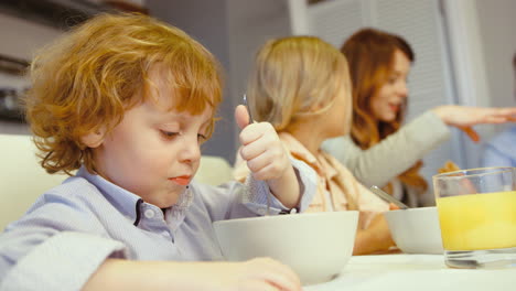 un niño encantador con ojos azules comiendo cereal mientras el resto de la familia habla en la cocina