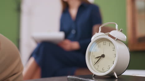 white timer resting on thick notebook atop table with blurred background of therapist seated on sofa holding pen and writing pad during thoughtful conversation in quiet therapy room