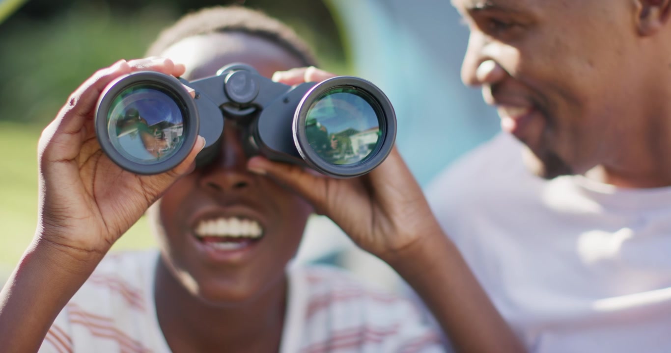 Padre e figlio africani che condividono un binocolo, il padre che lo guida mentre il figlio scruta per individuare una casa illuminata dal sole nel cortile.
