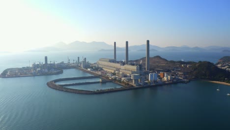 Drone-shot,-traveling-to-the-left,-of-a-big-coastal-coal-fired-power-plant-on-an-island-next-to-a-village-with-a-beach-during-a-beautiful-sunny-day