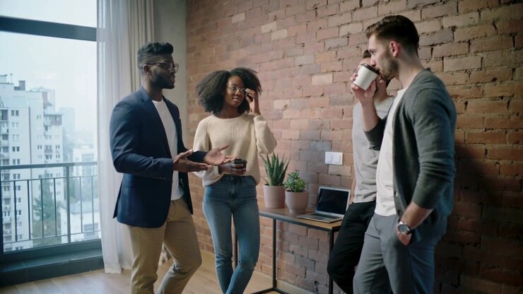L'uomo più a sinistra in blazer blu navy che inizia a parlare e spiega il progetto nell'area lounge dell'ufficio, con un portatile.