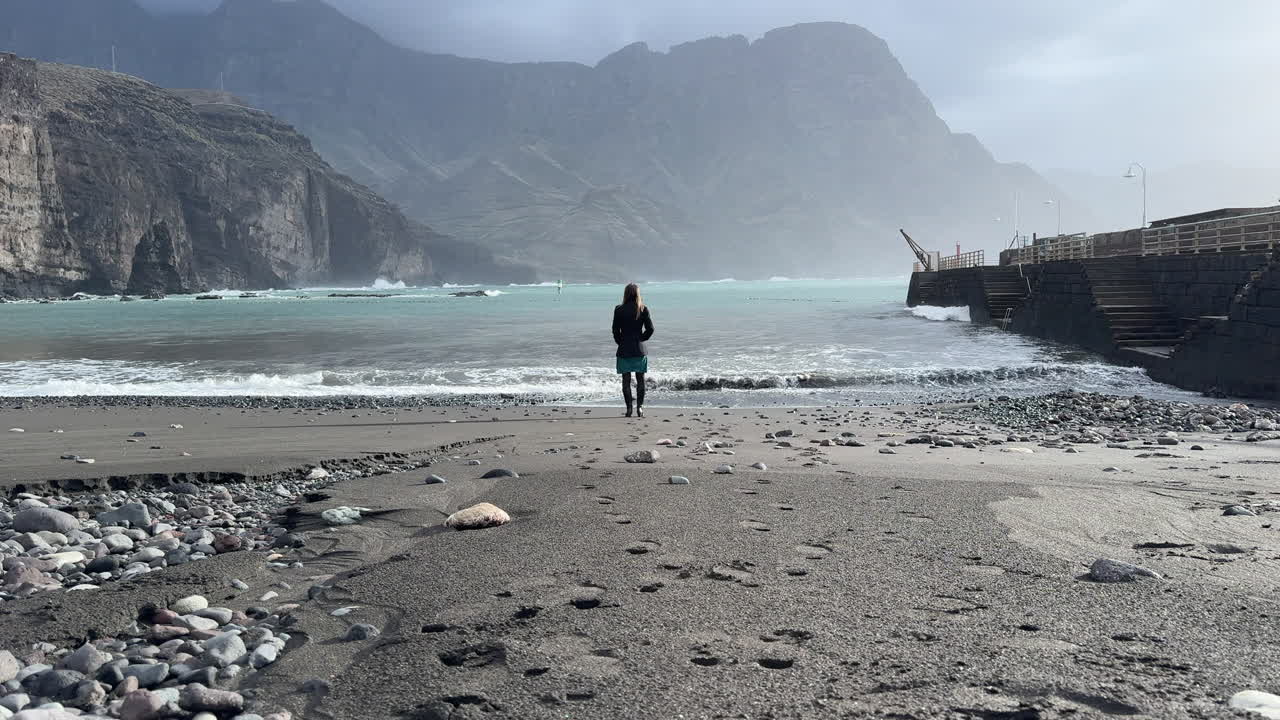 Donna matura con un cappotto che cammina su una spiaggia desolata verso un oceano mosso con scogliere sullo sfondo. Spiaggia di Agaete, Gran Canaria