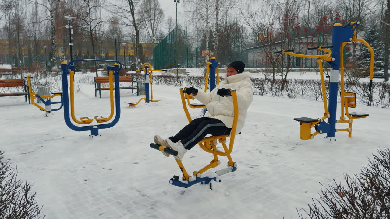Sports ground with diverse simulators in winter. Adult woman in white jacket training on snowing weather.