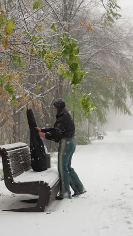 la señora quita el paquete de la guitarra y lo coloca en un banco cubierto de nieve en un parque de invierno sereno con árboles helados y una figura distante caminando por un camino nevado bajo la nieve suave que cae