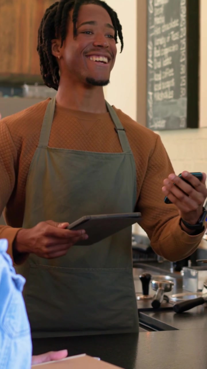 Vertical video: Taking payment, barista in olive apron making espresso and serving cups at counter
