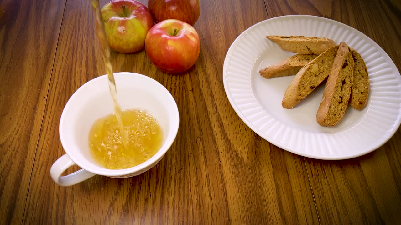 Slow motion shot of tea being poured into a cup with apples and cookies in the background